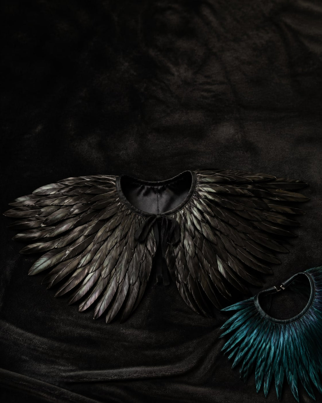 Overhead flat-lay of a black feather capelet opened out on dark velvet, feathers fanning outward from the neckline
