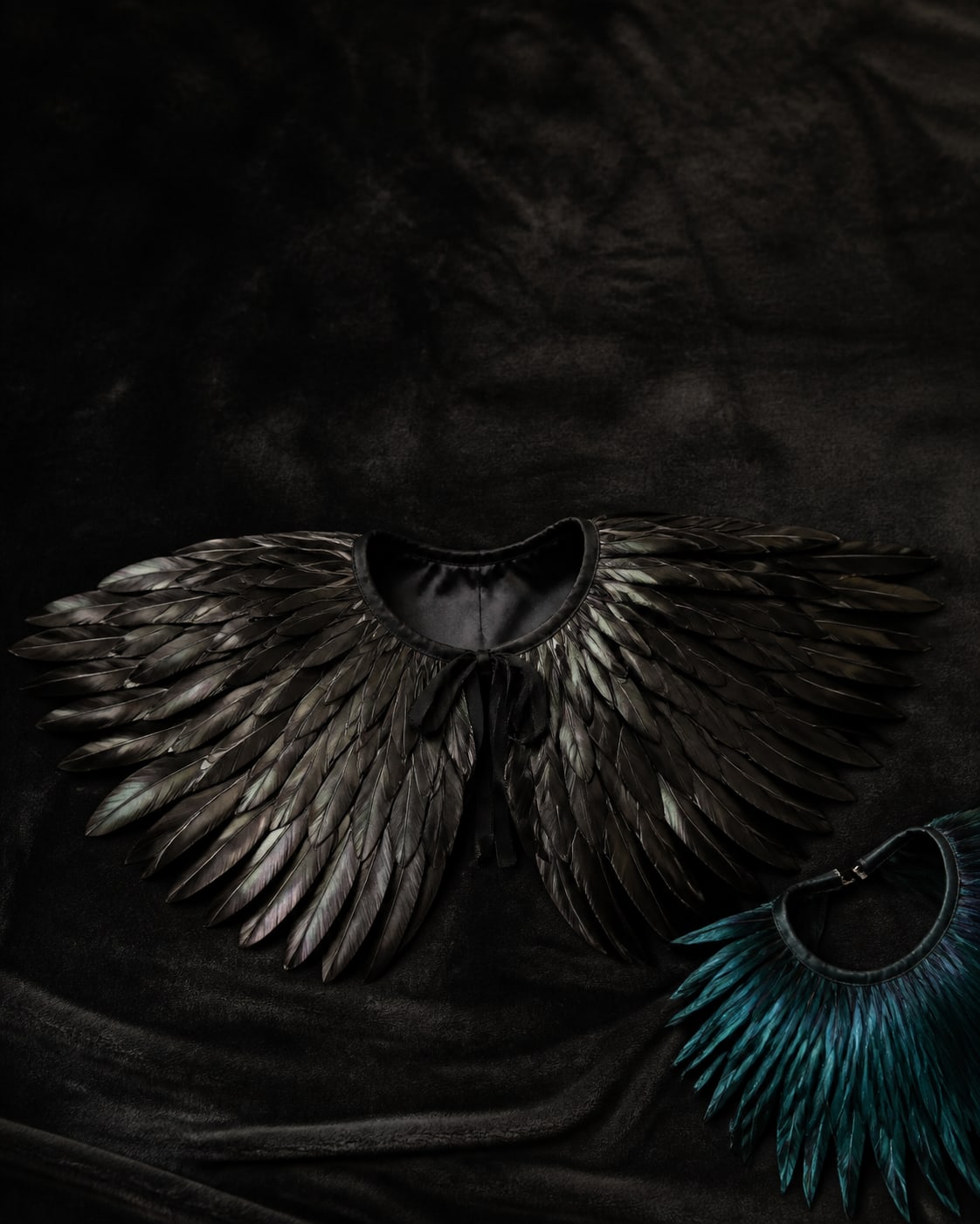 Overhead flat-lay of a black feather capelet opened out on dark velvet, feathers fanning outward from the neckline