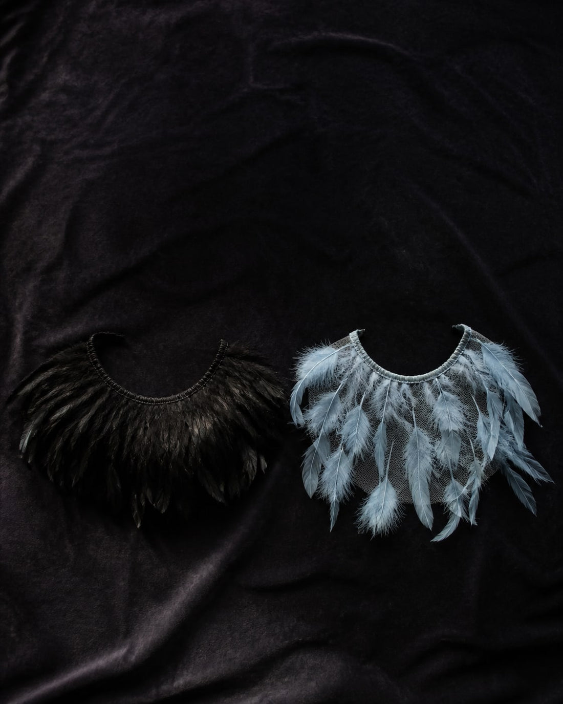 Overhead flat-lay comparing a dense opaque black feather collar with a see-through pale blue sheer mesh feather collar on dark velvet
