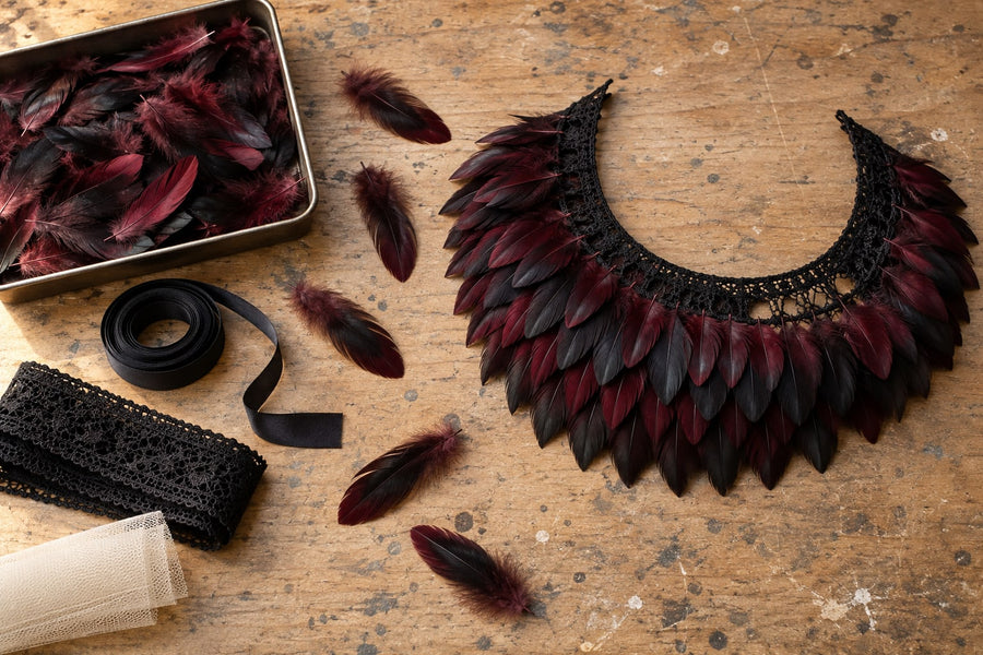 A workbench with a half-finished feather collar, the bare lace band still visible, beside loose dyed feathers, satin ribbon, lace edging and tulle