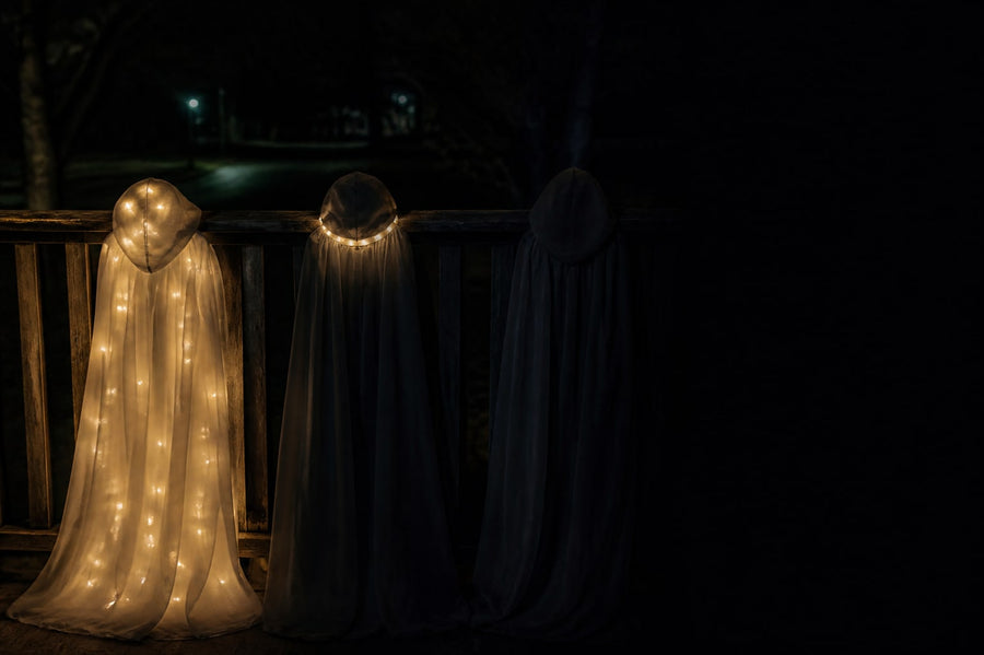 Three white hooded Halloween capes on a porch rail at night: one lit all over, one lit only at the collar, one unlit and nearly invisible