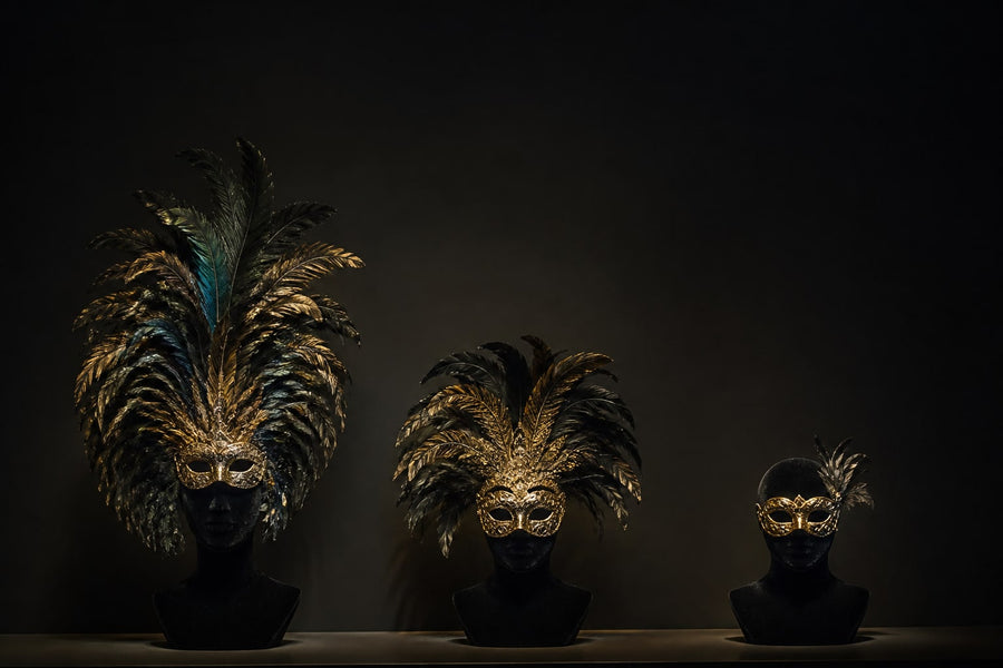 Three masquerade pieces on display busts: a full feather crown, a moderate plume, and a bare feathered eye-mask