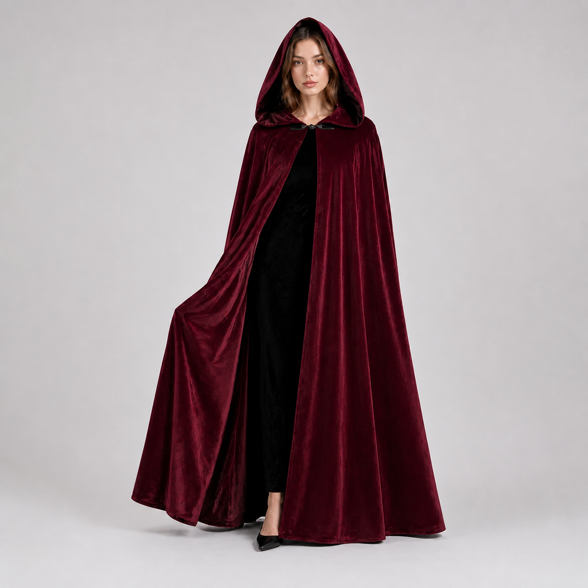 Full-body female model wearing a burgundy velvet-textured hooded cape with the hood up and one side held open