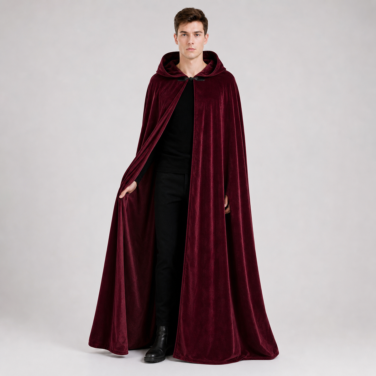 Full-body male model wearing a burgundy velvet-textured hooded cape over black clothing, with the front clasp fastened