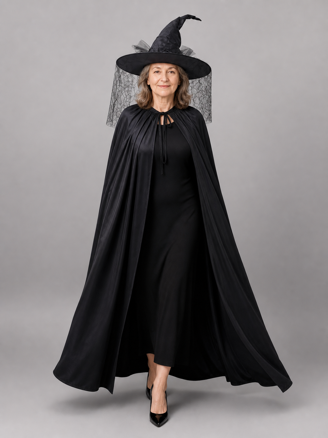 Black satin-look cloak with gathered neckline and pointed hat with mesh veil, shown full-body on a model