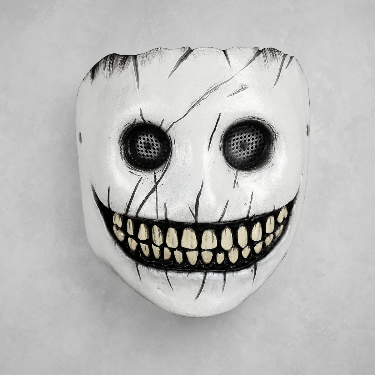 White full-face mask with black eye mesh, painted cracks, and a wide grin showing rows of yellowed teeth, shown against a gray background