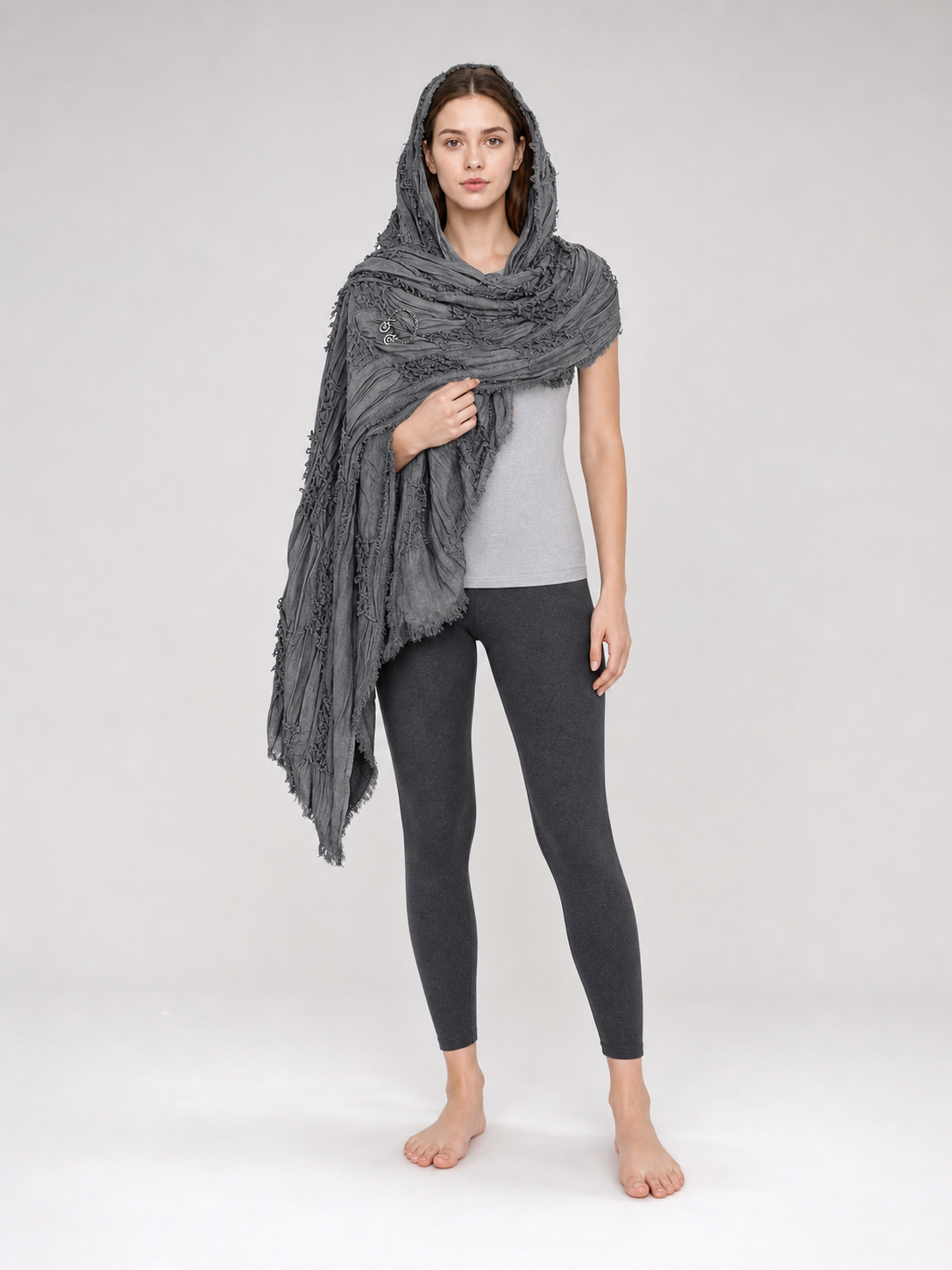 Gray textured scarf cape with hood and fringe, draped over a woman in a gray top and leggings in a studio
