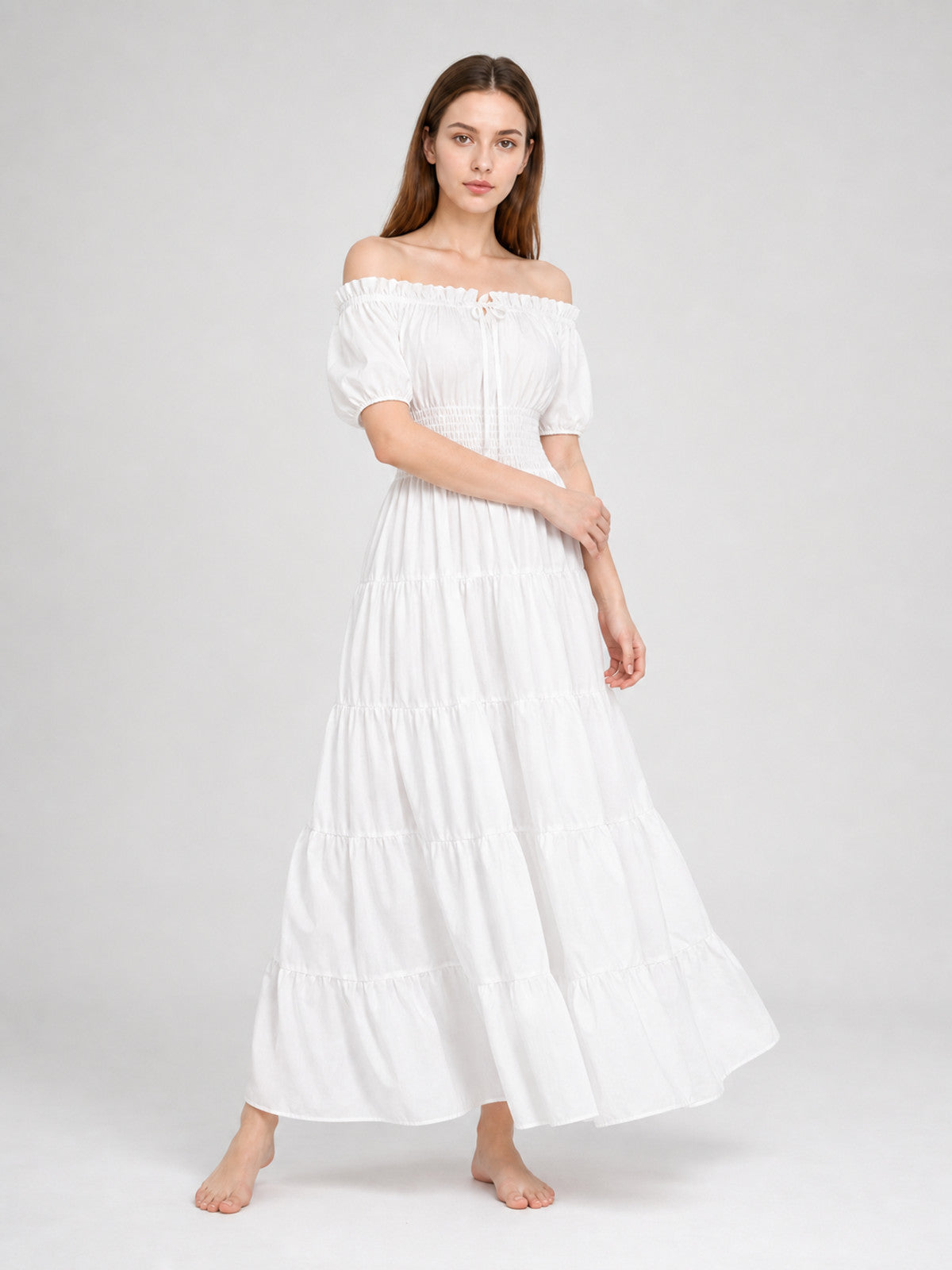 White off-shoulder maxi dress shown on a walking model, with short puff sleeves, a gathered waist, and a flowing tiered skirt