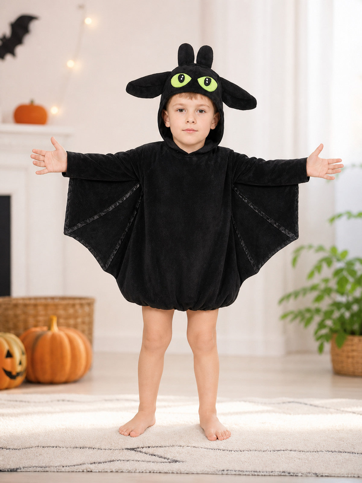 Black plush bat costume shown full-body on a child in a furnished room, with hood, ears, eyes, and wing panels
