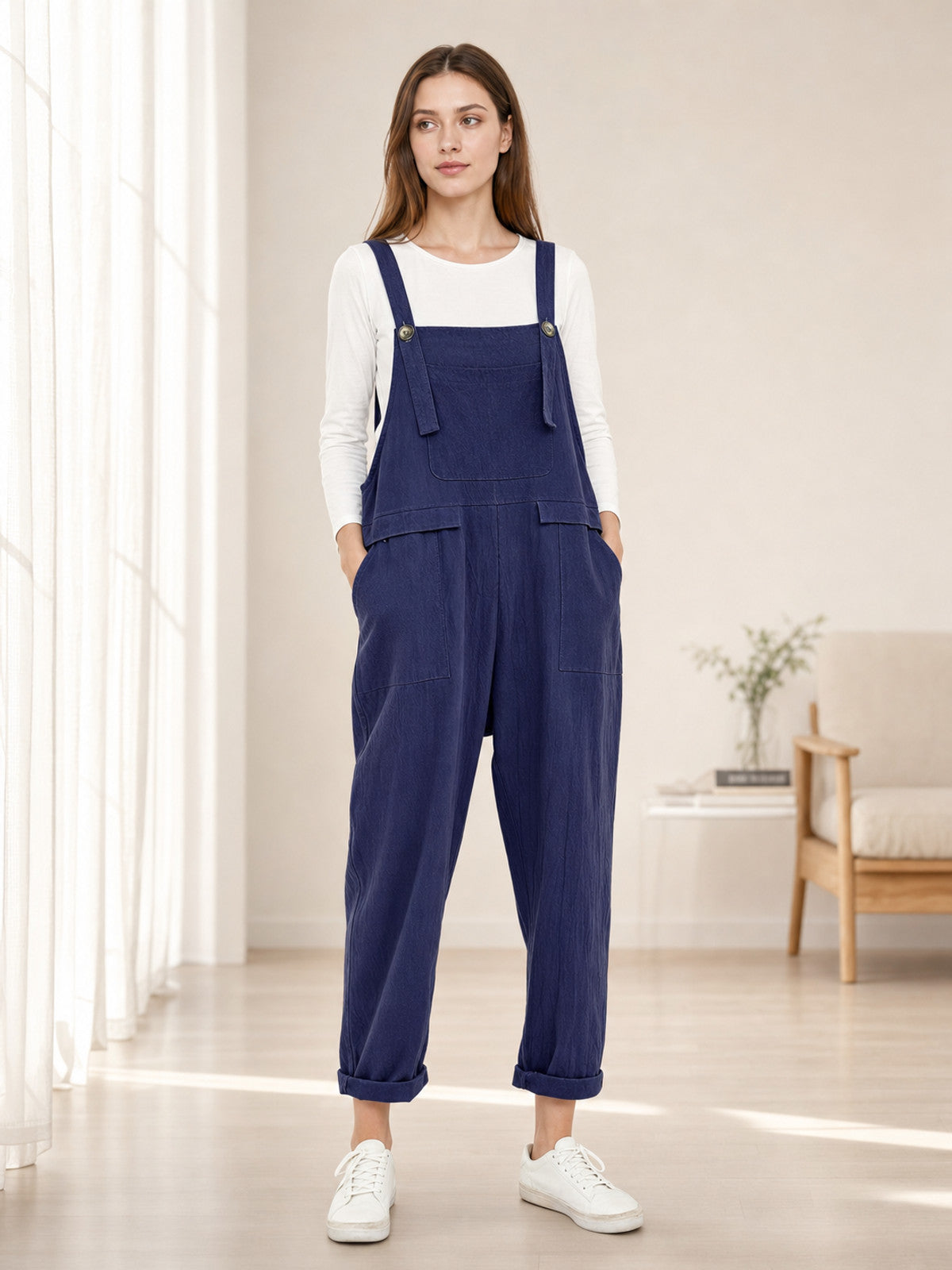 Navy blue bib overalls modeled in a bright room over a white long-sleeve shirt, showing front pockets, loose legs, and cuffed hems