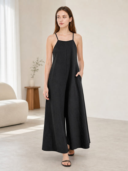 Black wide-leg jumpsuit with thin straps and side pockets, modeled in a bright room with a sofa and wooden side table