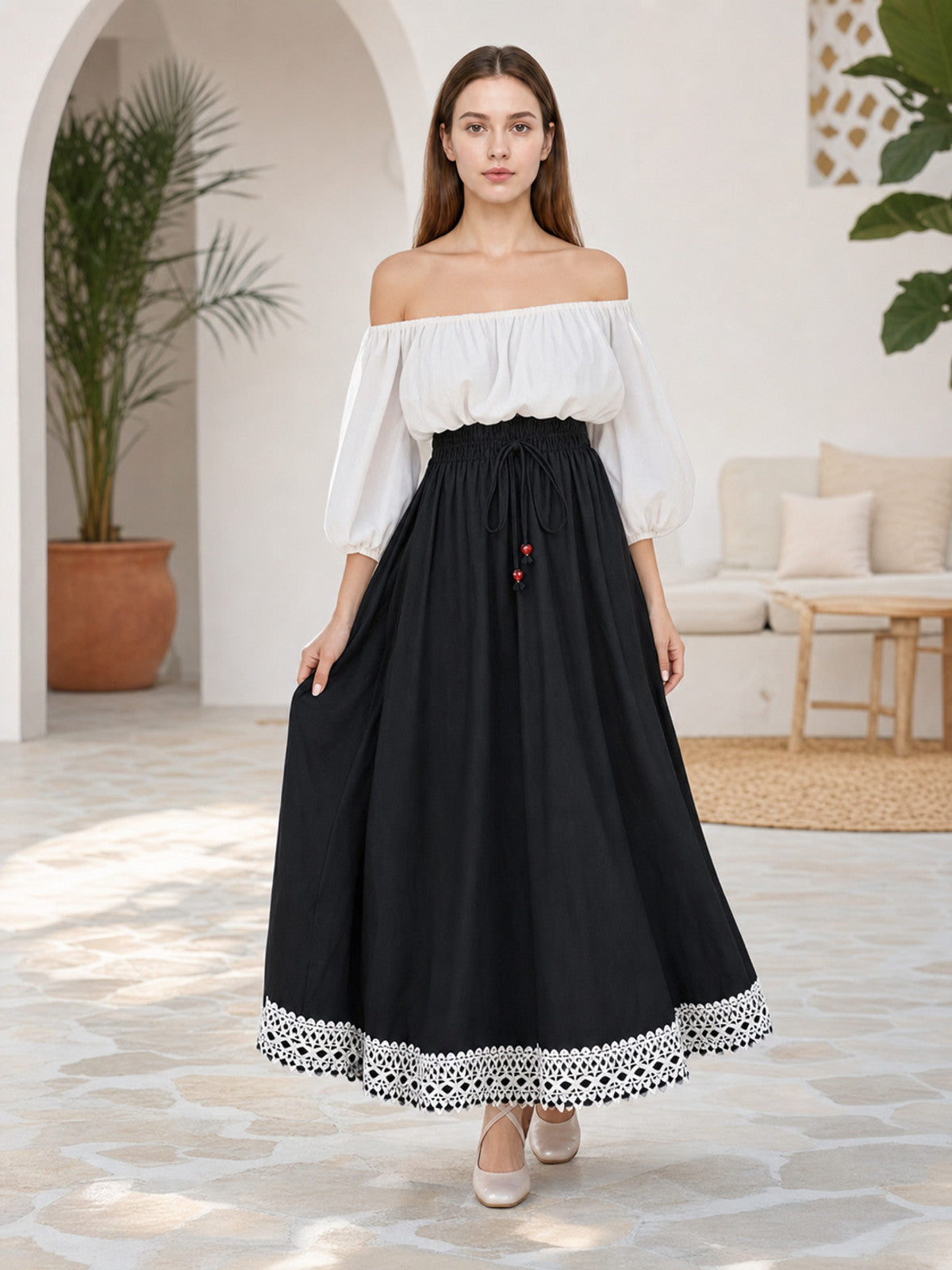 Full-body model wearing a flowing black maxi skirt with a gathered waist, red bead drawstrings, and white lace hem trim in a living room