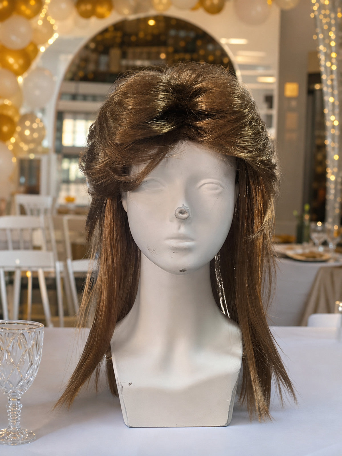 Brown costume wig with a full swept top and long side sections, displayed on a mannequin in a decorated indoor setting