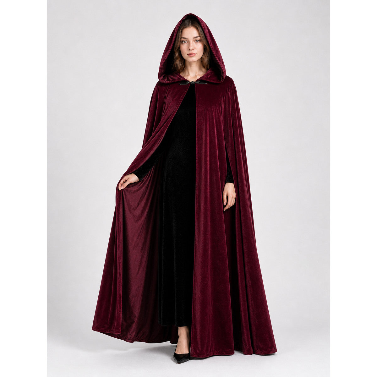 Full-body female model wearing a burgundy velvet-textured hooded cape, holding the open front to show the black outfit underneath