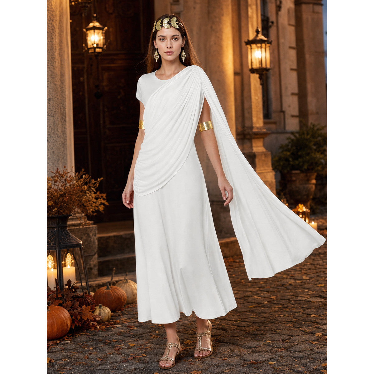 Full-body model wearing a white draped dress with a flowing train in an outdoor courtyard, styled with gold accessories and sandals