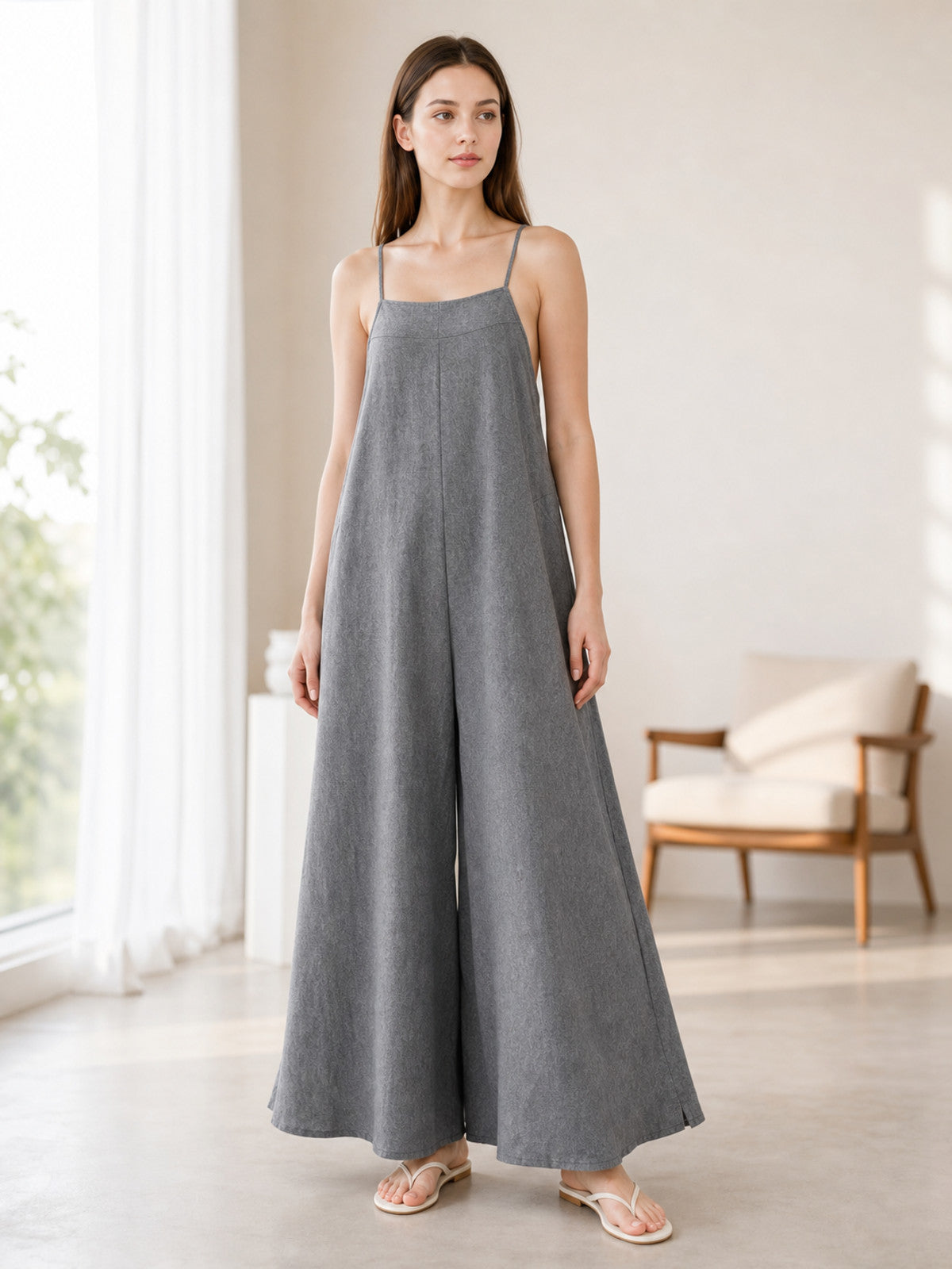 Gray wide-leg jumpsuit with a softly textured finish and thin shoulder straps, shown full-length on a model in a bright room