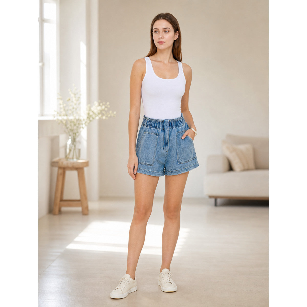 Light blue denim shorts shown full-body on a model in a living room, styled with a white tank top and sneakers