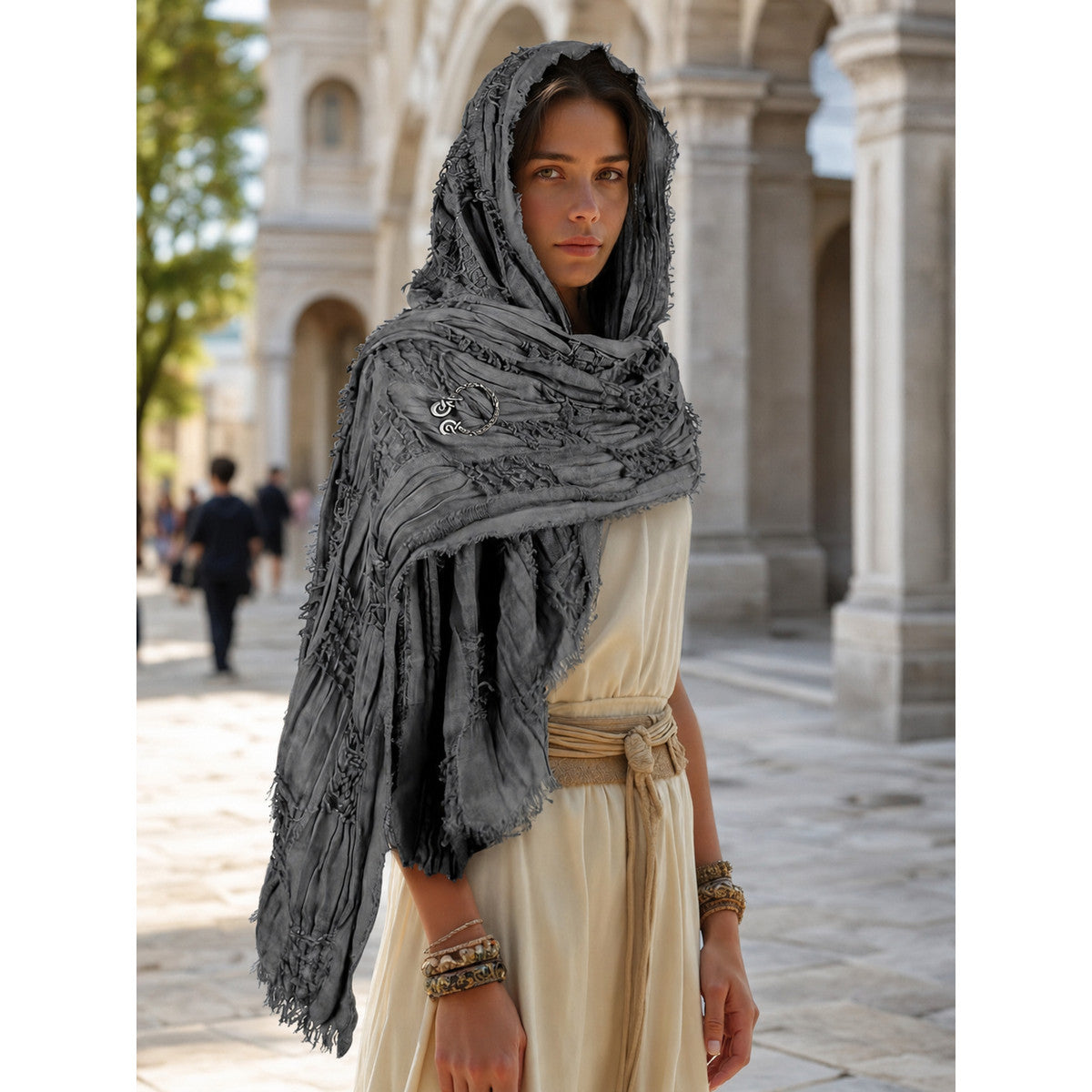 Gray fringed hooded scarf cape with a metal clasp, worn over a cream outfit in an outdoor colonnade