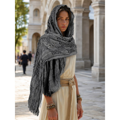 Gray fringed hooded scarf cape with a metal clasp, worn over a cream outfit in an outdoor colonnade