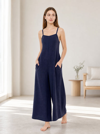 Navy blue wide-leg jumpsuit with thin straps and side pockets shown on a barefoot model in a bright living room