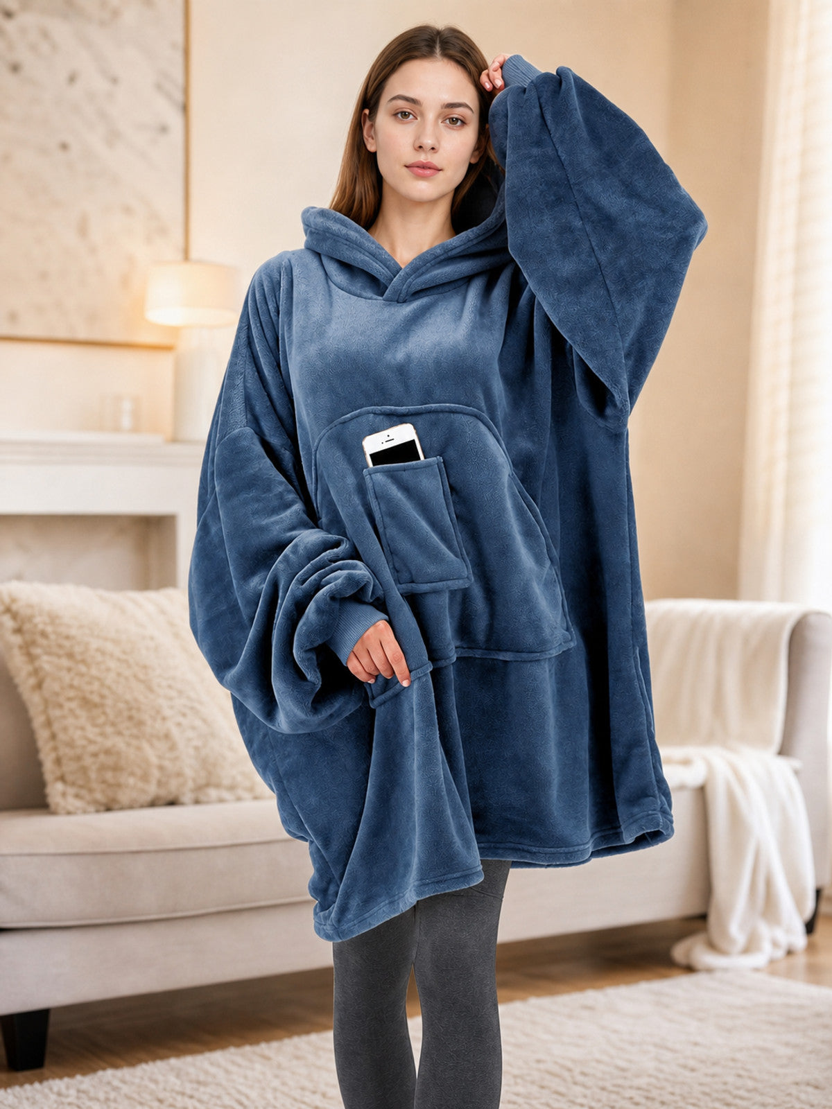 Blue plush fleece hooded blanket hoodie shown on a model in a living room, with oversized sleeves and a front pouch pocket