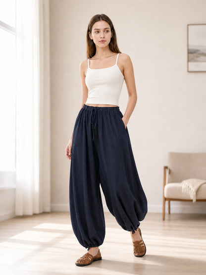 Dark blue pants styled on a model in a bright room, showing the elastic waist, side pockets, wide legs, and gathered hems
