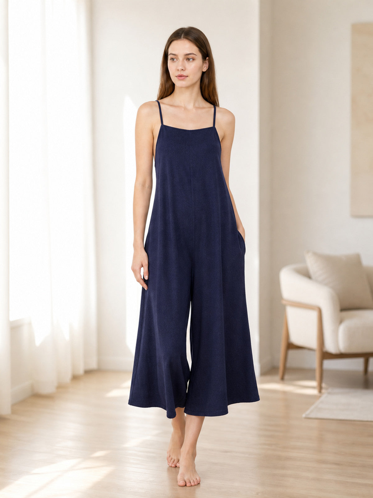 Dark blue wide-leg jumpsuit with thin straps and side pockets, shown full-length on a barefoot model in a bright room