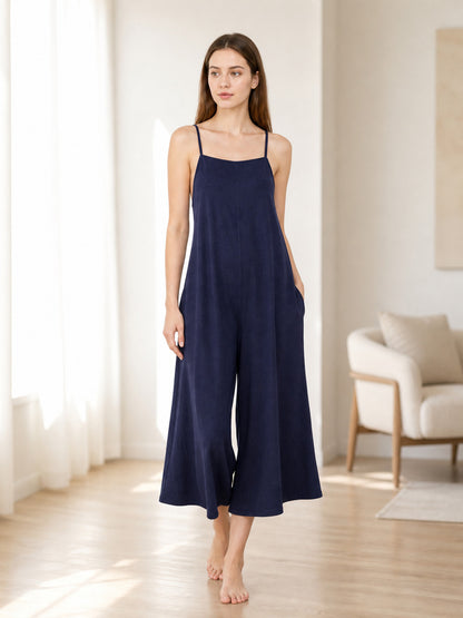 Dark blue wide-leg jumpsuit with thin straps and side pockets, shown full-length on a barefoot model in a bright room