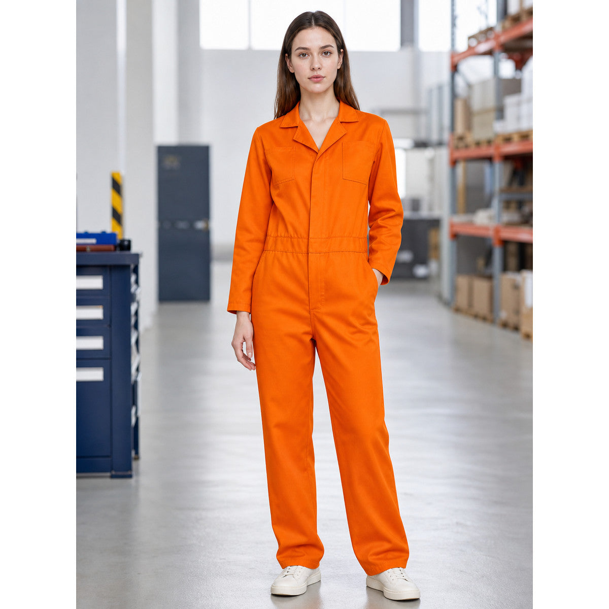 Woman in an orange long-sleeve jumpsuit standing in a bright warehouse, with a notched collar, chest pockets, elastic waist, and white sneakers