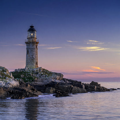 Scenic photograph of a stone lighthouse on rocky coast beside calm water at sunset