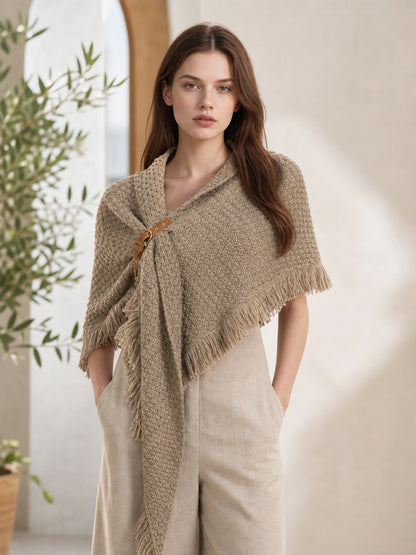 Beige textured cape styled on a model with light trousers, featuring a side clasp, pointed front panel, and fringed trim