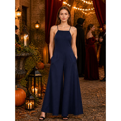 Navy blue wide-leg jumpsuit shown full-length on a model, with thin straps and side pockets in a lantern-lit setting