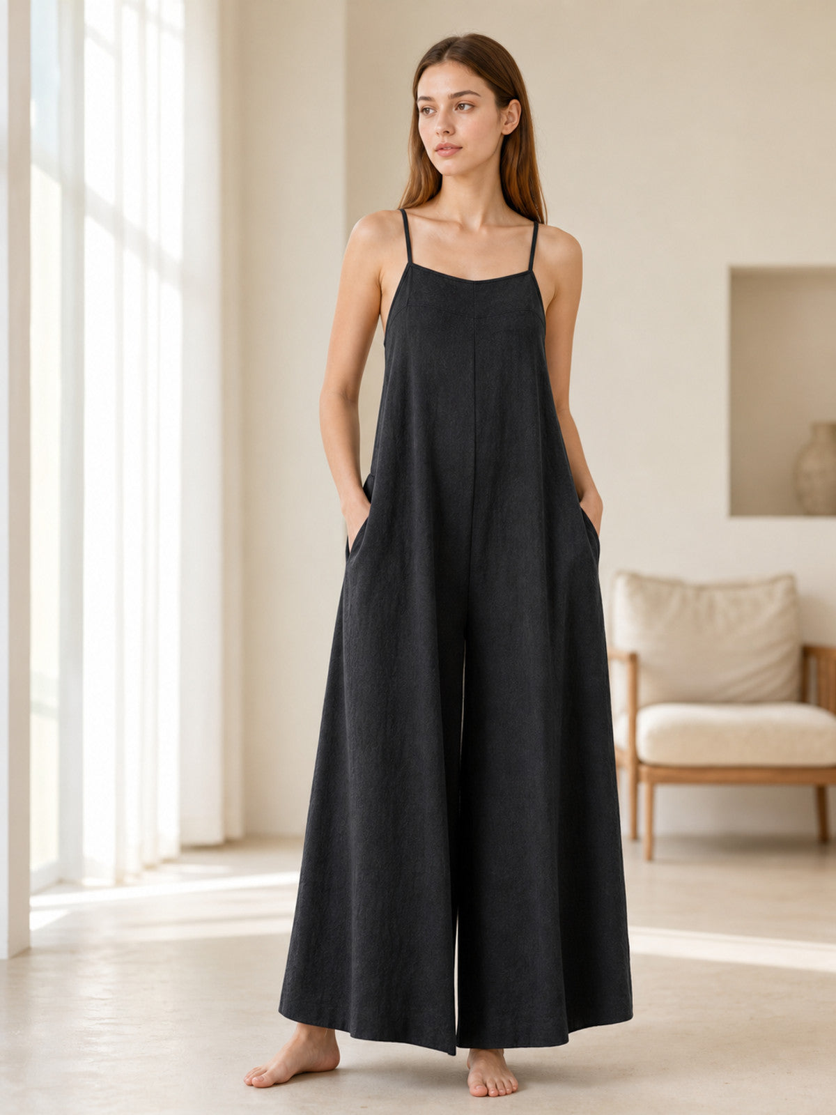 Black wide-leg jumpsuit with thin straps and side pockets, shown full-body on a model in a bright living room