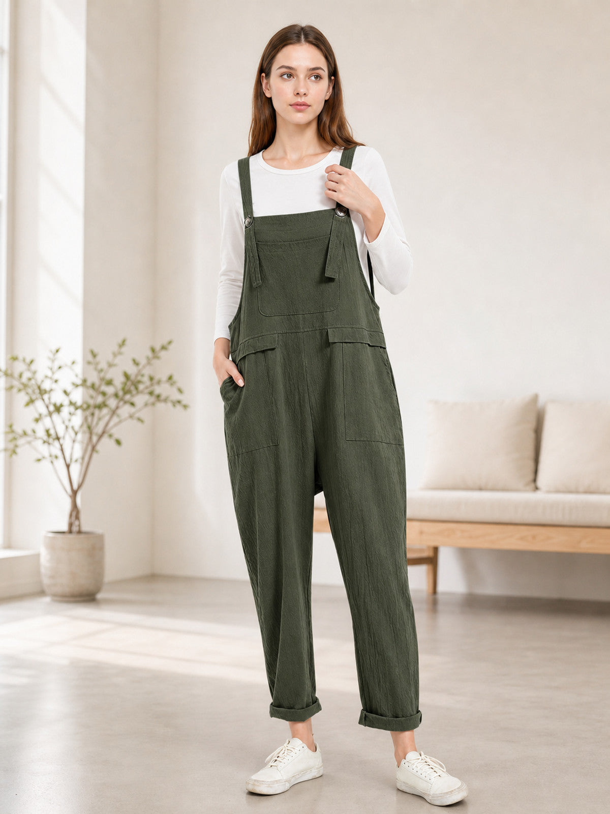 Full-body model wearing dark green bib overalls with a chest pocket, large side pockets, rolled cuffs, and white sneakers in a living room