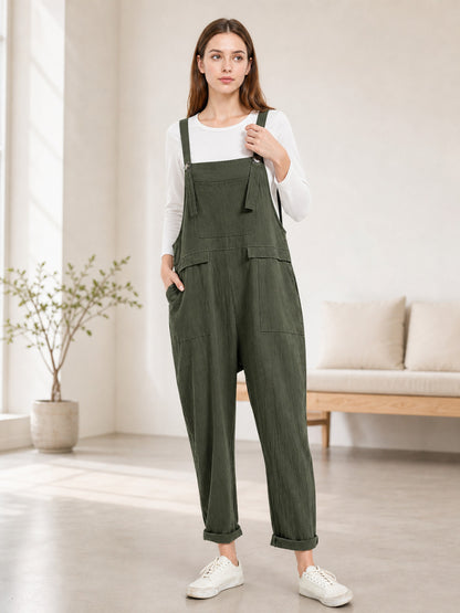 Full-body model wearing dark green bib overalls with a chest pocket, large side pockets, rolled cuffs, and white sneakers in a living room