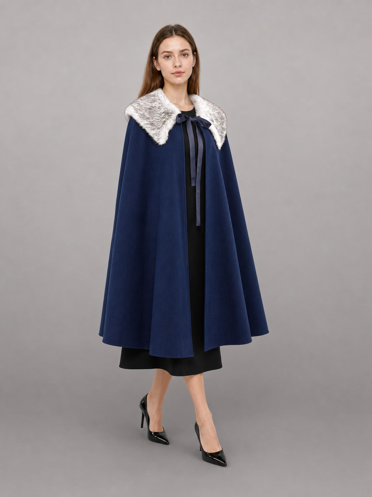 Navy blue cape with a white faux fur collar and front ribbon tie, shown on a model in a full-length studio portrait