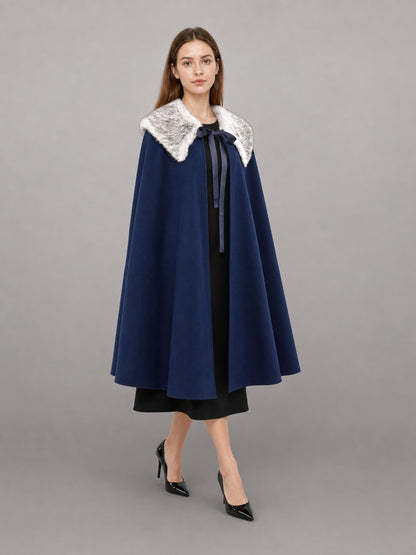Navy blue cape with a white faux fur collar and front ribbon tie, shown on a model in a full-length studio portrait