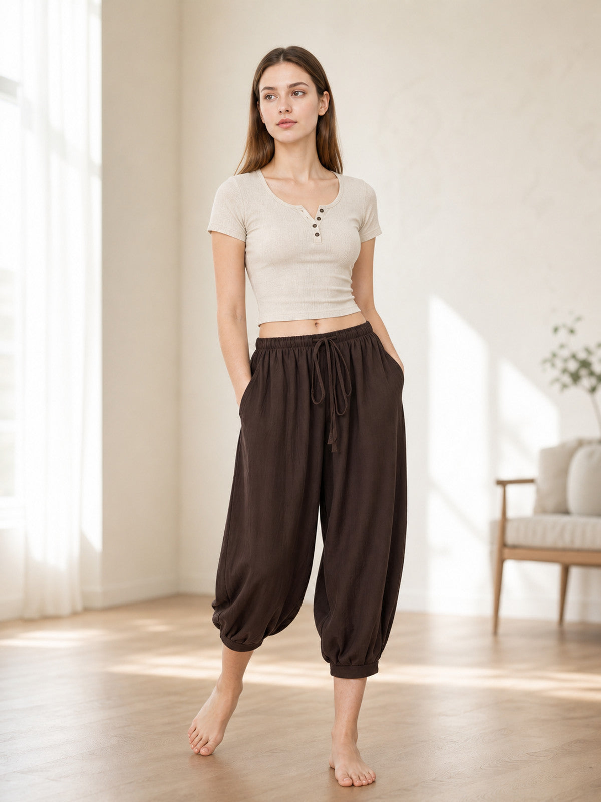 Brown cropped harem pants styled on a standing model indoors, showing the elastic waist, drawstring, side pockets and gathered cuffs