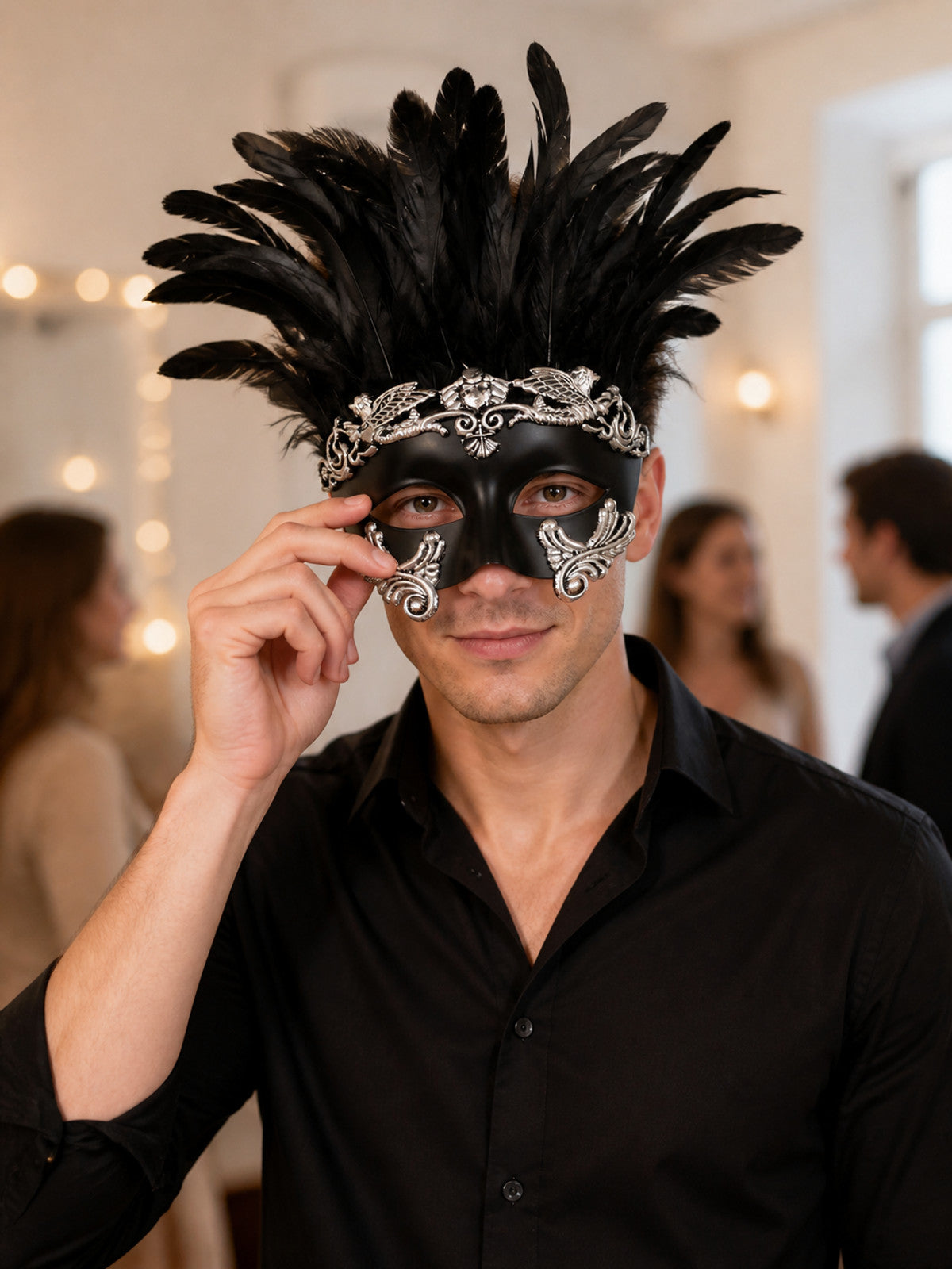 Man wearing a black feather masquerade mask with silver detailing at an indoor gathering, adjusting the mask with one hand