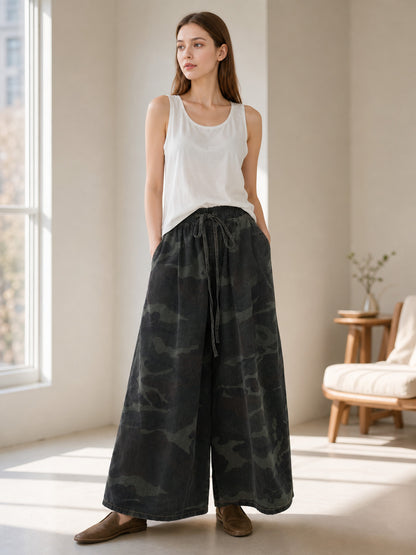 Dark camouflage wide-leg cropped jeans shown on a model indoors, paired with a white tank top and brown flats