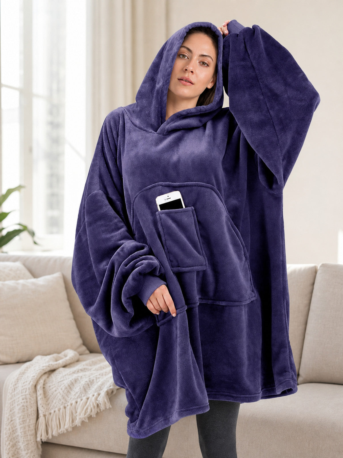 Dark purple plush hooded blanket poncho worn by a model indoors, showing oversized sleeves, hood, and front pocket