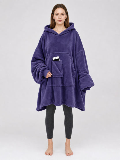 Dark purple plush hooded blanket poncho shown full-body on a model, with oversized sleeves and a front kangaroo pocket holding a phone