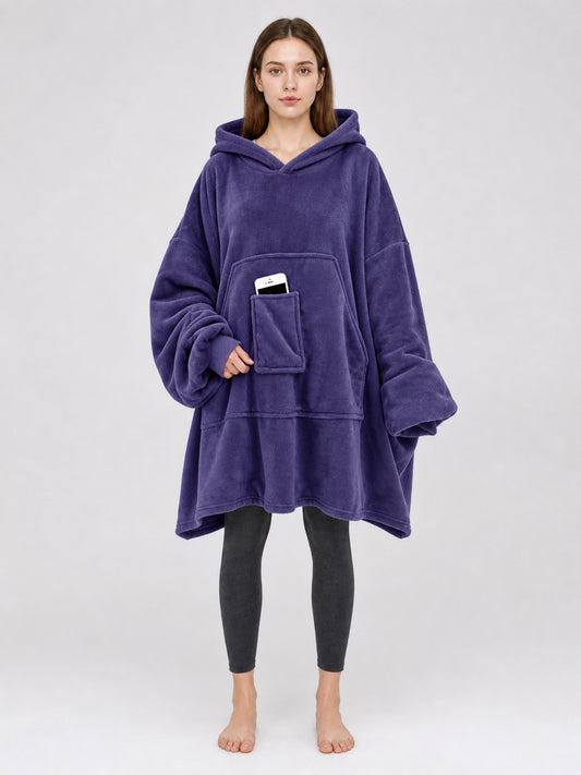 Dark purple plush hooded blanket poncho shown full-body on a model, with oversized sleeves and a front kangaroo pocket holding a phone