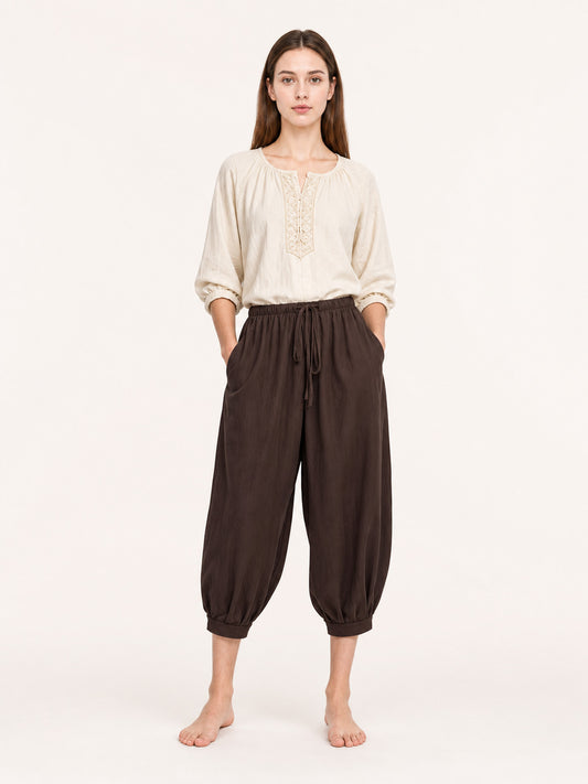 Brown cropped harem pants on a full-body model, with an elastic waist, front drawstring, side pockets and gathered ankle cuffs