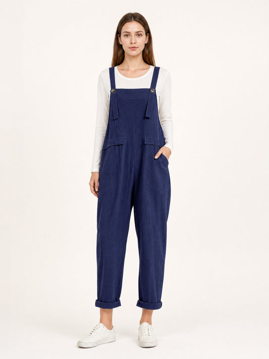 Navy blue bib overalls modeled full-body over a white long-sleeve shirt, with shoulder straps, front pockets, tapered legs, and cuffed hems