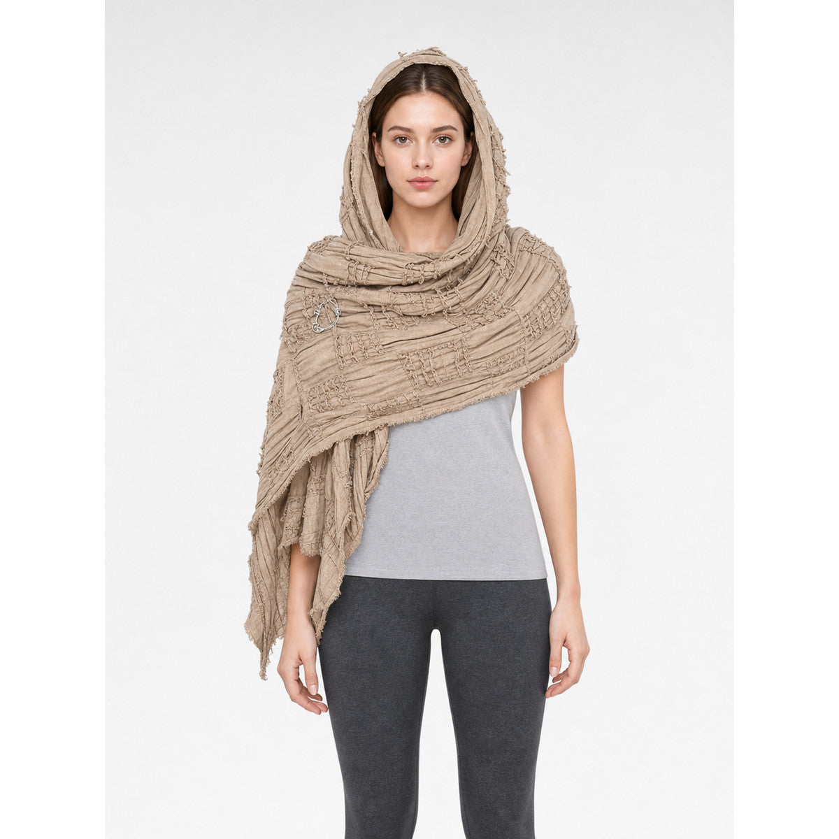 Khaki hooded scarf cape with textured woven bands and fringed edges, worn over a gray shirt and dark leggings