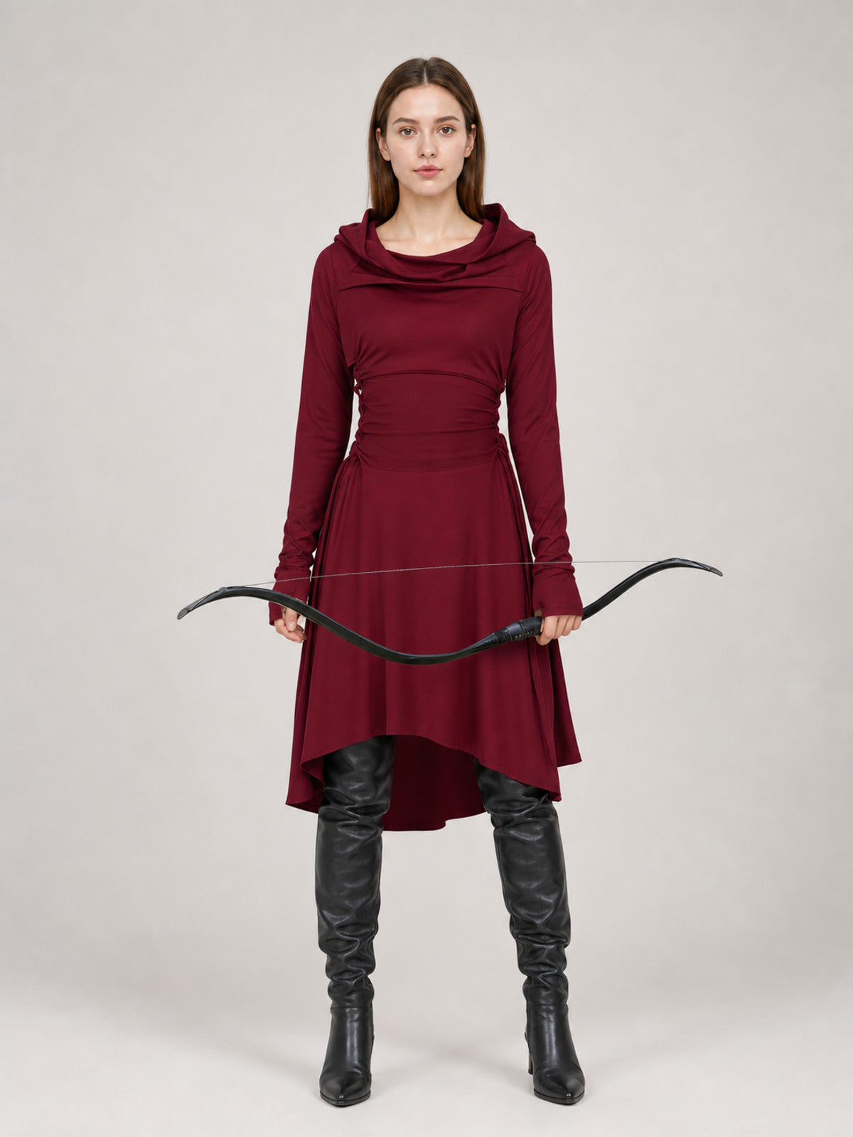 Burgundy hooded tunic in smooth fabric, shown full-body on a model with long sleeves, draped neckline, uneven hem, black boots, and a bow