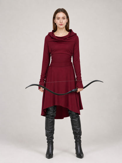 Burgundy hooded tunic in smooth fabric, shown full-body on a model with long sleeves, draped neckline, uneven hem, black boots, and a bow