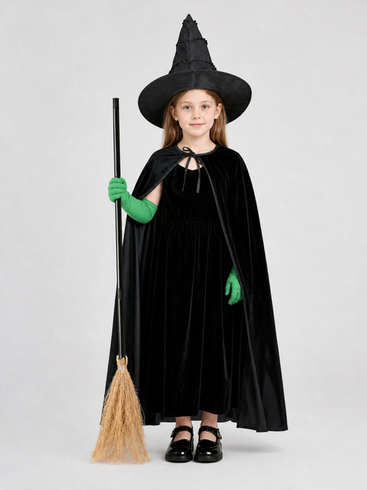 Full-body child model wearing a black velvet dress and cape, pointed hat, green gloves, and holding a broom