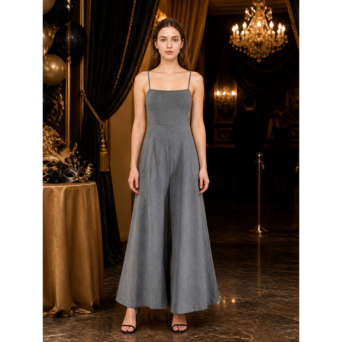 Gray wide-leg jumpsuit with thin shoulder straps and a front center pleat, shown full-length on a model in an indoor evening setting